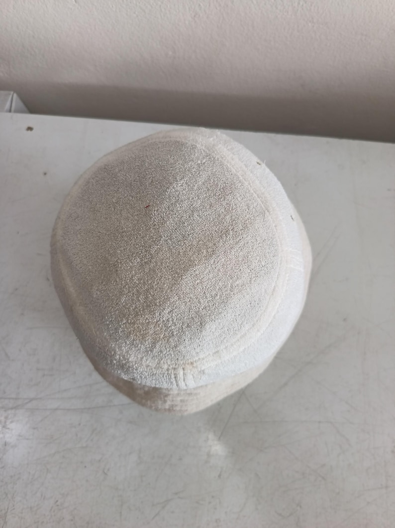 Pode incluir: Um gorro redondo de tecido texturizado em tom esbranqui&ccedil;ado, com topo plano e uma faixa lateral estruturada. O material tem uma apar&ecirc;ncia macia e ligeiramente felpuda, com costuras vis&iacute;veis que definem a coroa circular e o painel lateral.