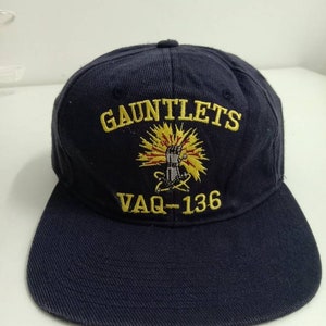 May include: A black and yellow baseball cap with the text "GAUNTLETS VAQ-136" embroidered on the front. The cap features a yellow fist with lightning bolts and a red star.