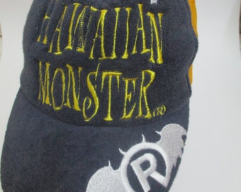 Può includere: Un cappello da baseball blu e giallo con il testo "Hawaiian Monster" ricamato in giallo. Il cappello ha un pipistrello bianco con un simbolo di marchio registrato ricamato sul lato.