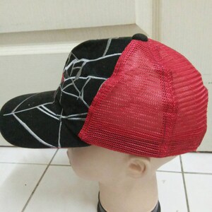 May include: A black and red baseball cap with a white spider web design. The back of the cap is made of red mesh.