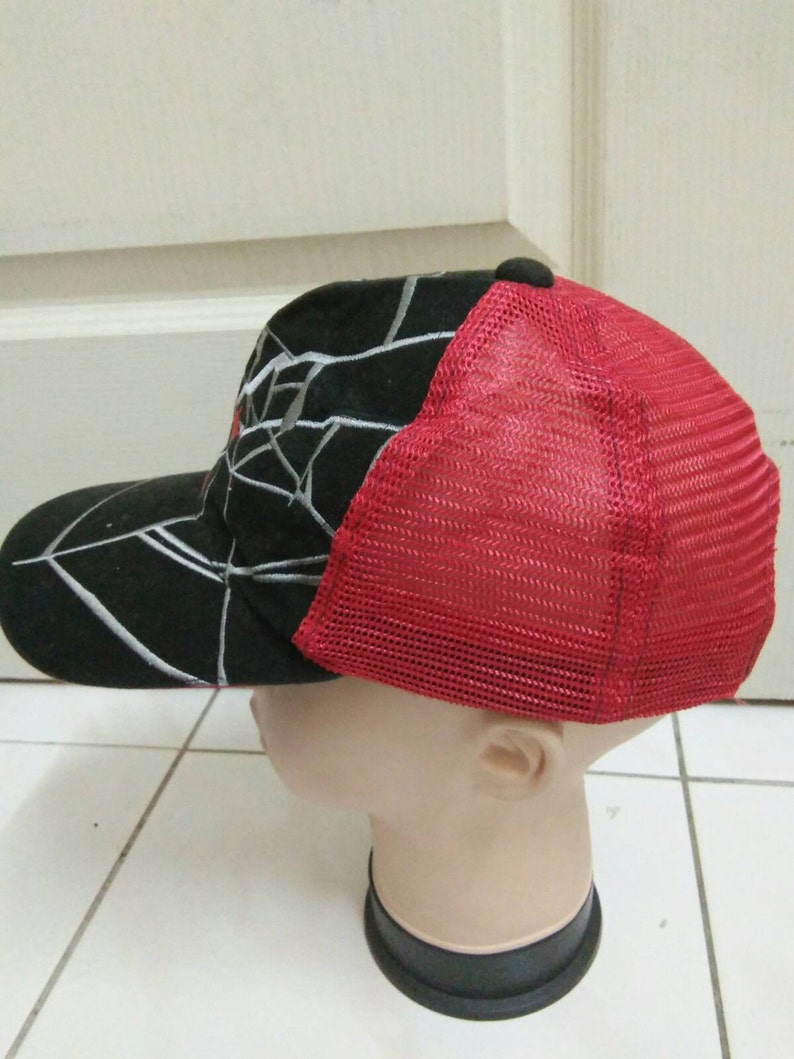 May include: A black and red baseball cap with a white spider web design. The back of the cap is made of red mesh.
