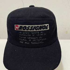 May include: A black cap with a flat brim featuring the brand name "ROSSIGNOL" in white and red. The cap has embroidered text in French. The cap is made of a dark fabric.