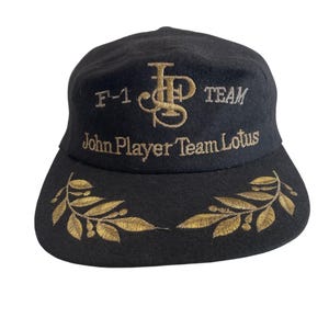 May include: Black cap with gold embroidered text and leaf design. The text reads "F-1 TEAM John Player Team Lotus". The cap has a curved brim and a classic, sporty design.