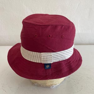 May include: A burgundy bucket hat with a white and gray striped band. The hat has a small blue tag with a logo. The hat is displayed on a mannequin head.