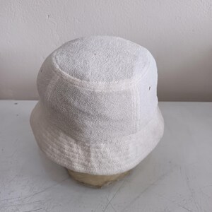 Pode incluir: Um chap&eacute;u bucket branco s&oacute;lido feito de tecido felpudo texturizado. O chap&eacute;u apresenta uma copa arredondada e uma aba inclinada para baixo que circunda a cabe&ccedil;a. O material tem uma superf&iacute;cie macia de la&ccedil;os, t&iacute;pica de tecidos atoalhados.