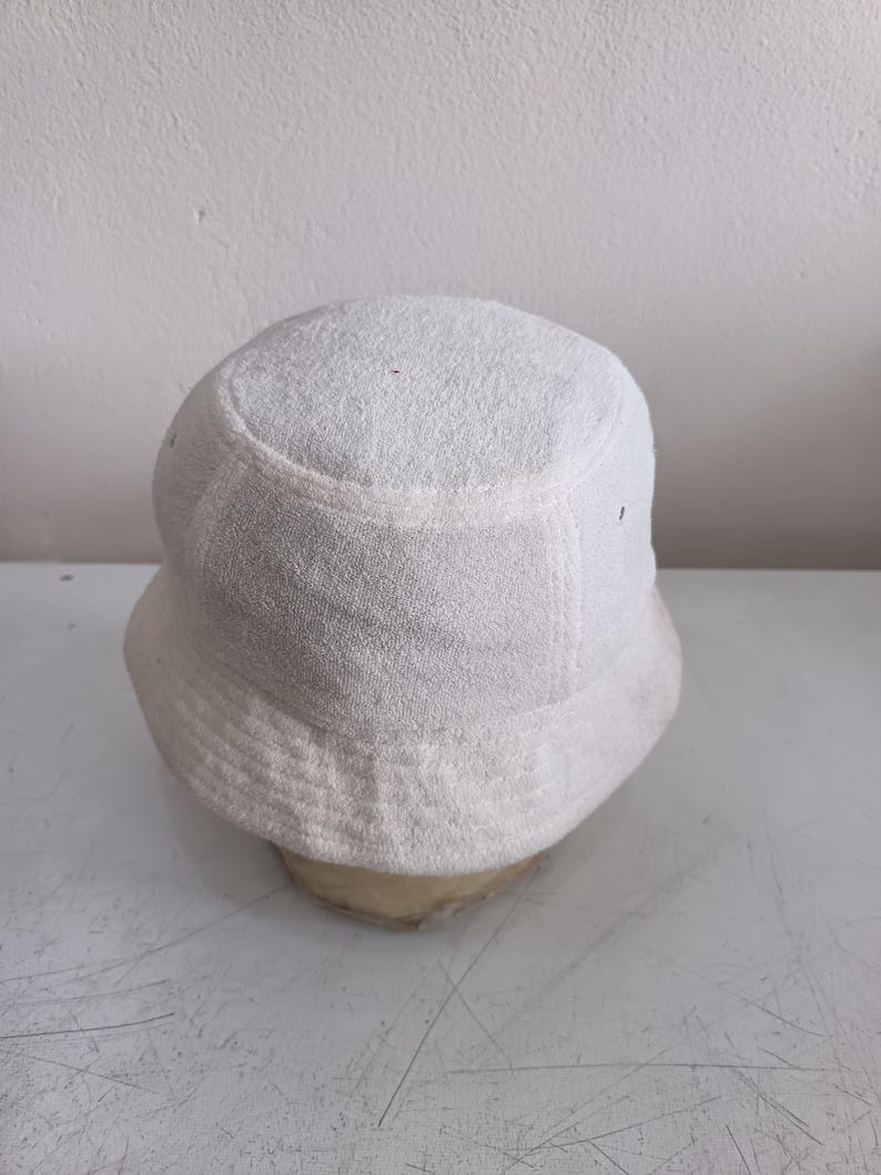 Pode incluir: Um chap&eacute;u bucket branco s&oacute;lido feito de tecido felpudo texturizado. O chap&eacute;u apresenta uma copa arredondada e uma aba inclinada para baixo que circunda a cabe&ccedil;a. O material tem uma superf&iacute;cie macia de la&ccedil;os, t&iacute;pica de tecidos atoalhados.