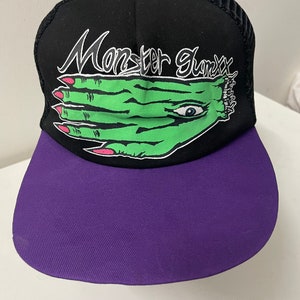 May include: Black and purple trucker hat with a green hand with an eye and the text "Monster Glamxx" in white.