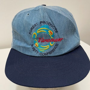 May include: A blue denim baseball cap with a black brim. The front of the cap features an embroidered logo that says "Forest Products Safety Conference Vancouver" in a circular design with yellow, blue, and red accents.