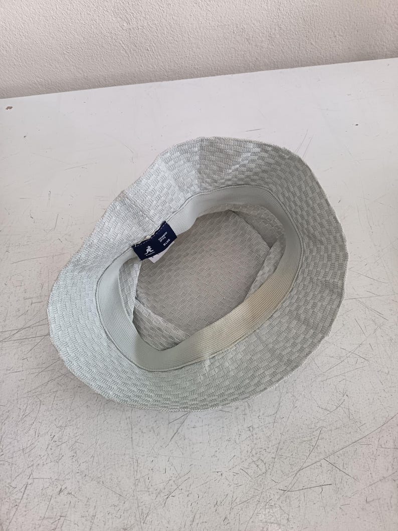 May include: A light gray bucket hat with a textured, woven pattern. The interior features a white sweatband and a dark blue Kangol brand label with a kangaroo logo and the text "KANGOL" and "BLUE" printed in white on the inner rim.