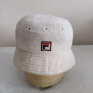 Pode incluir: Um chap&eacute;u bucket de tecido felpudo branco com um pequeno emblema quadrado na frente contendo o log&oacute;tipo da Fila em vermelho, azul-marinho e branco. O chap&eacute;u tem uma copa plana, aba inclinada com costuras e ilh&oacute;s de ventila&ccedil;&atilde;o.