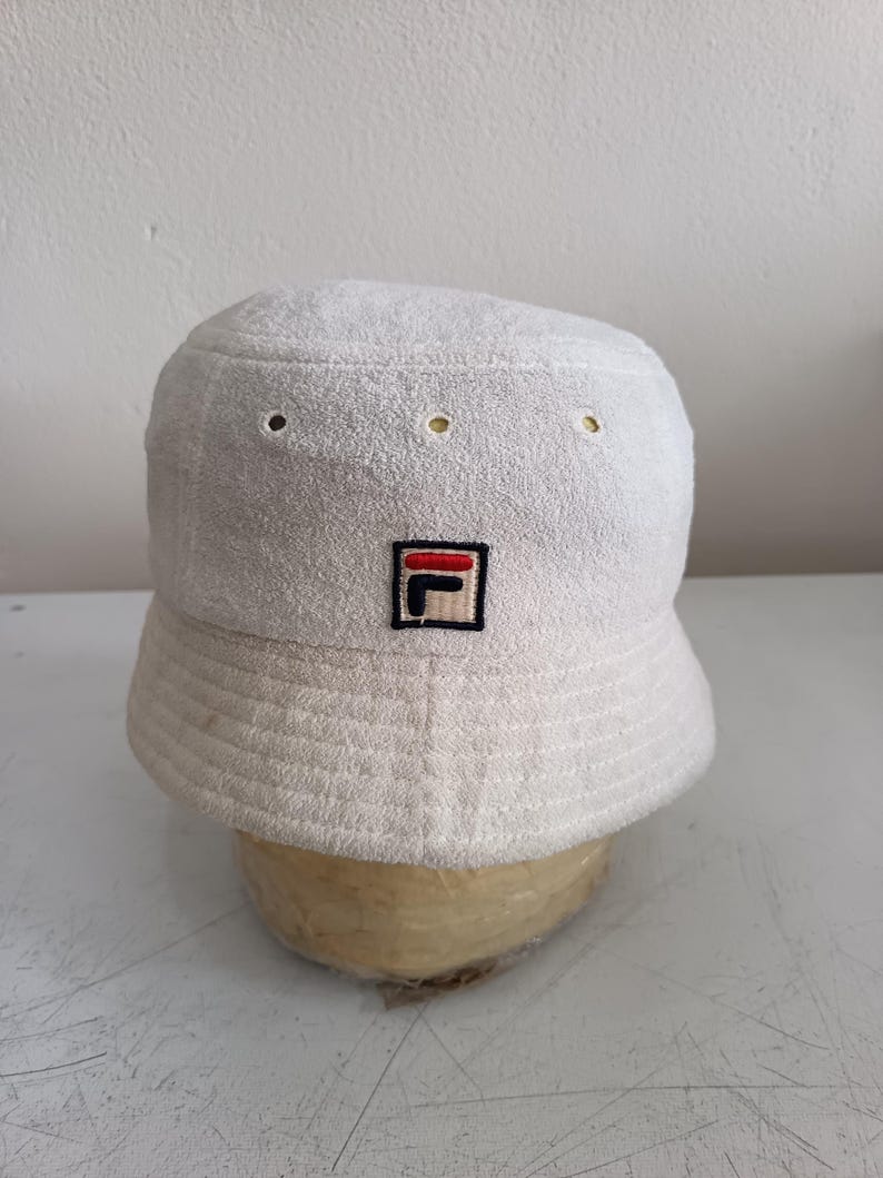 Pode incluir: Um chap&eacute;u bucket de tecido felpudo branco com um pequeno emblema quadrado na frente contendo o log&oacute;tipo da Fila em vermelho, azul-marinho e branco. O chap&eacute;u tem uma copa plana, aba inclinada com costuras e ilh&oacute;s de ventila&ccedil;&atilde;o.