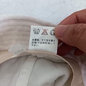 Pode incluir: Um chap&eacute;u branco com uma etiqueta de cuidados contendo texto em japon&ecirc;s e s&iacute;mbolos de lavagem. A etiqueta inclui &iacute;cones para lavagem &agrave; m&atilde;o a 30 graus, sem branqueamento e secagem &agrave; sombra. O texto diz: 洗濯後、手で型を整え日陰で自然乾燥して下さい。