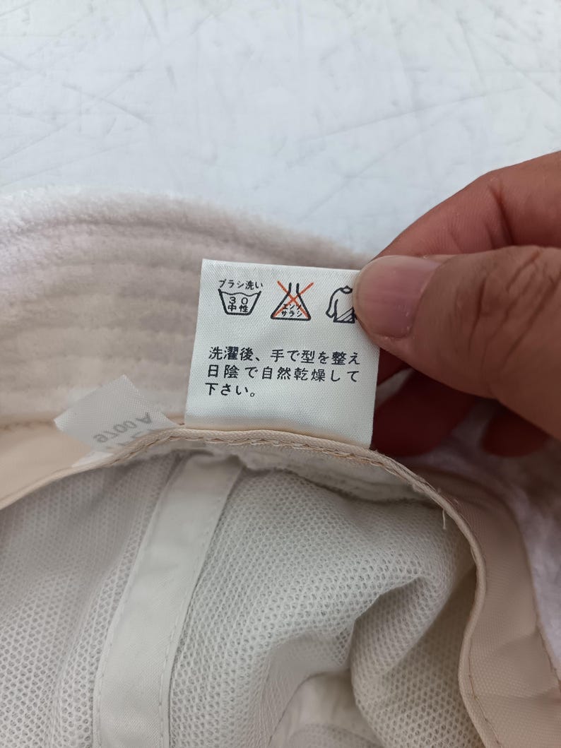 Pode incluir: Um chap&eacute;u branco com uma etiqueta de cuidados contendo texto em japon&ecirc;s e s&iacute;mbolos de lavagem. A etiqueta inclui &iacute;cones para lavagem &agrave; m&atilde;o a 30 graus, sem branqueamento e secagem &agrave; sombra. O texto diz: 洗濯後、手で型を整え日陰で自然乾燥して下さい。