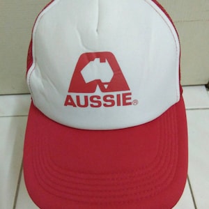 Könnte beinhalten: Eine rot-weiße Trucker-Kappe mit einem roten Logo einer stilisierten Karte von Australien und dem Wort "Aussie" in weißen Buchstaben. Die Kappe hat ein Netzrücken.