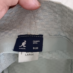 May include: A light blue bucket hat with a textured weave pattern. Inside, a dark blue rectangular label features the white Kangol kangaroo logo, the text FOUNDED 38.83, and the word BLUE. A white tag below displays the size LARGE.