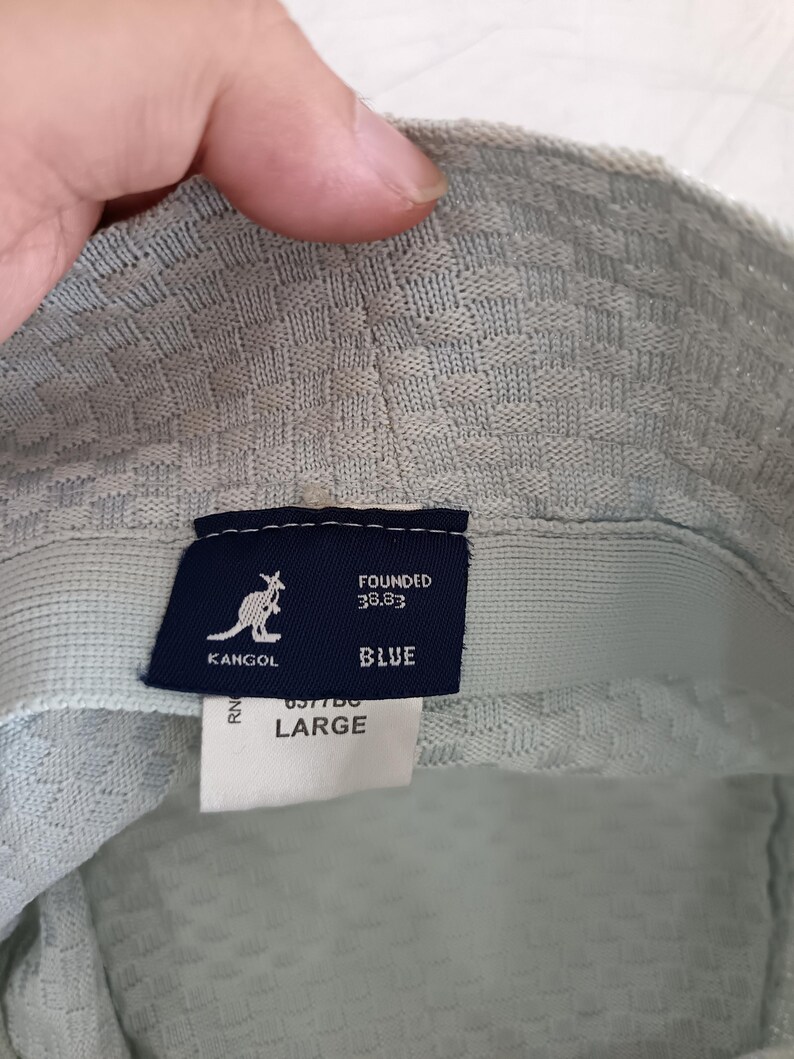 May include: A light blue bucket hat with a textured weave pattern. Inside, a dark blue rectangular label features the white Kangol kangaroo logo, the text FOUNDED 38.83, and the word BLUE. A white tag below displays the size LARGE.