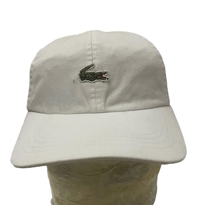 May include: A white cotton baseball cap with a green crocodile logo on the front. The logo has a red mouth and a pink tongue.