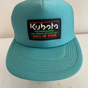 Peut inclure: Casquette de camionneur turquoise avec une visière incurvée et un dos en filet. L'avant présente un écusson rectangulaire avec le logo Kubota et le texte "THE PROFESSIONAL HAS HUMAN TECHNOLOGY. HALL OF FAME."