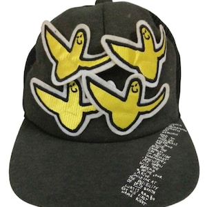 May include: A black mesh baseball cap with three yellow bird patches. The patches have white outlines and smiling faces. The cap has a black brim and a black crown. The text on the brim reads: "ENCH REPOLYAN THIS LES MISTRIBUALS THE JANE GET THE TWICE SIDE BY SIDE CRAAY IN ARSULUT HARMONEY EXCUSE WHEN BREAD THE ARIVE ORIVE, GUR SMILE ING AT THE POSIBILITY OF DETH DEATH GET HIS FIRST CURTIS RAMBO NAME RIGHT"