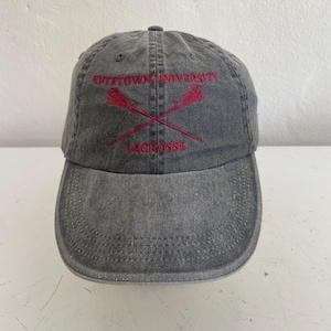 May include: A gray Kutztown University Lacrosse baseball cap. The cap features red embroidered text and crossed lacrosse sticks. The hat has a curved brim and an adjustable strap.