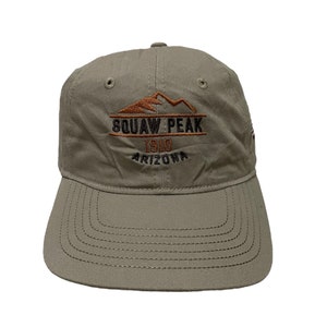 May include: A khaki-colored baseball cap with the text "SQUAW PEAK 1910 ARIZONA" embroidered in brown and black. The cap features a mountain graphic above the text and a curved brim.