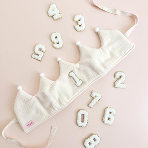 May include: A white crown with gold numbers 1 and 2 on it. The crown is made of soft fabric and has a tie to secure it. There are ten white numbers with gold outlines scattered around the crown, from 0 to 9.