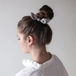 May include: A woman with long brown hair styled in a messy bun with a gray bow hair tie. She is wearing a white shirt with a ruffled collar.
