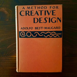 May include: A vintage book titled "A Method for Creative Design" by Adolfo Best-Maugard. The book has a peach-colored cover with black text and design elements. The book is set against a dark wood background.
