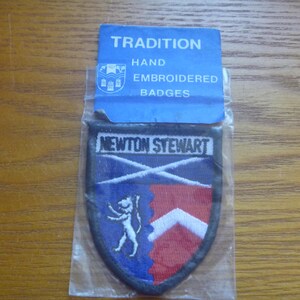 May include: A blue and red embroidered patch with the text "NEWTON STEWART" and a white cross on a blue background. The patch is packaged in a plastic bag with a blue label that reads "TRADITION HAND EMBROIDERED BADGES".