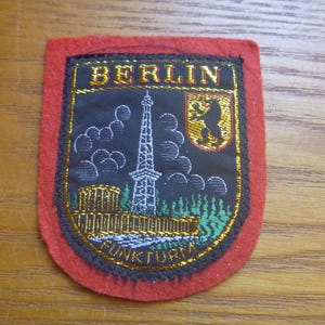 May include: A red, black and gold embroidered patch with the word "BERLIN" in gold lettering. The patch features a black silhouette of the Berlin Television Tower, a black silhouette of the Berlin bear, and the words "FUNKTURM" in gold lettering.