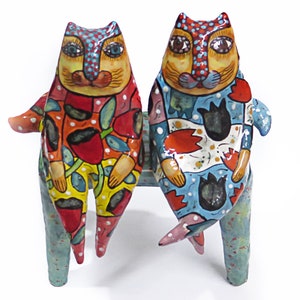 May include: Two colourful ceramic cats sitting on a blue bench. The cats are decorated with a variety of patterns, including flowers, dots and stripes. The bench is also decorated with a blue and white pattern.