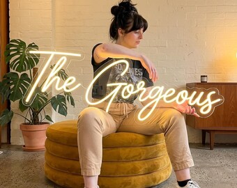 May include: A neon sign that reads "The Gorgeous" in a cursive font. The sign is lit up with a warm white light. The sign is being held by a person sitting on a yellow ottoman. The person is wearing a black t-shirt and tan pants.