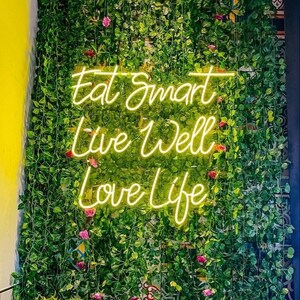 May include: A yellow neon sign with the text Eat Smart, Live Well, Love Life mounted on a wall covered in dense green artificial ivy vines with small pink flower accents. The sign features a cursive script font and emits a bright glow.