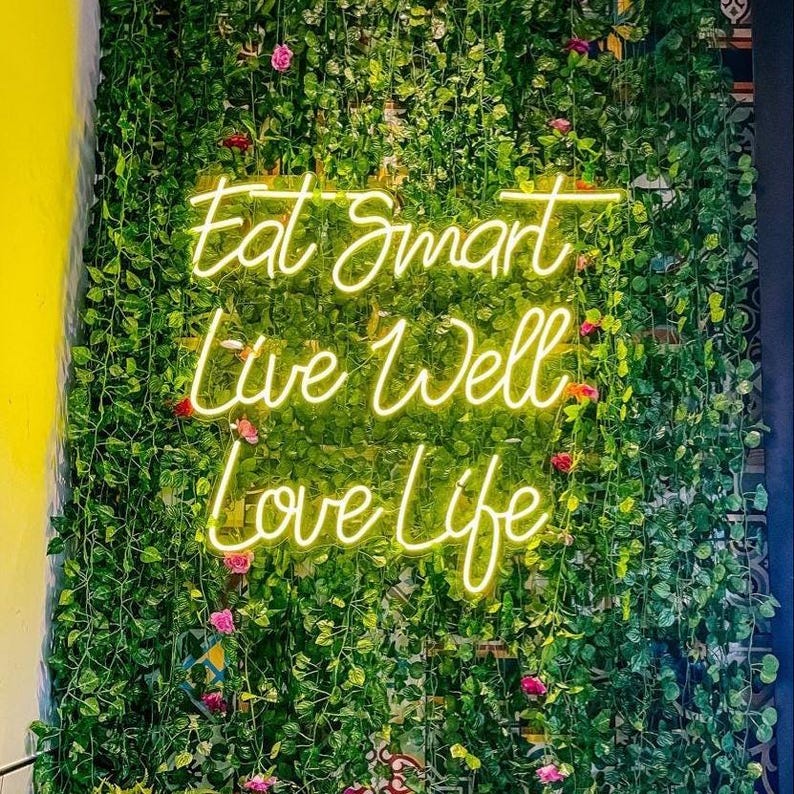May include: A yellow neon sign with the text Eat Smart, Live Well, Love Life mounted on a wall covered in dense green artificial ivy vines with small pink flower accents. The sign features a cursive script font and emits a bright glow.