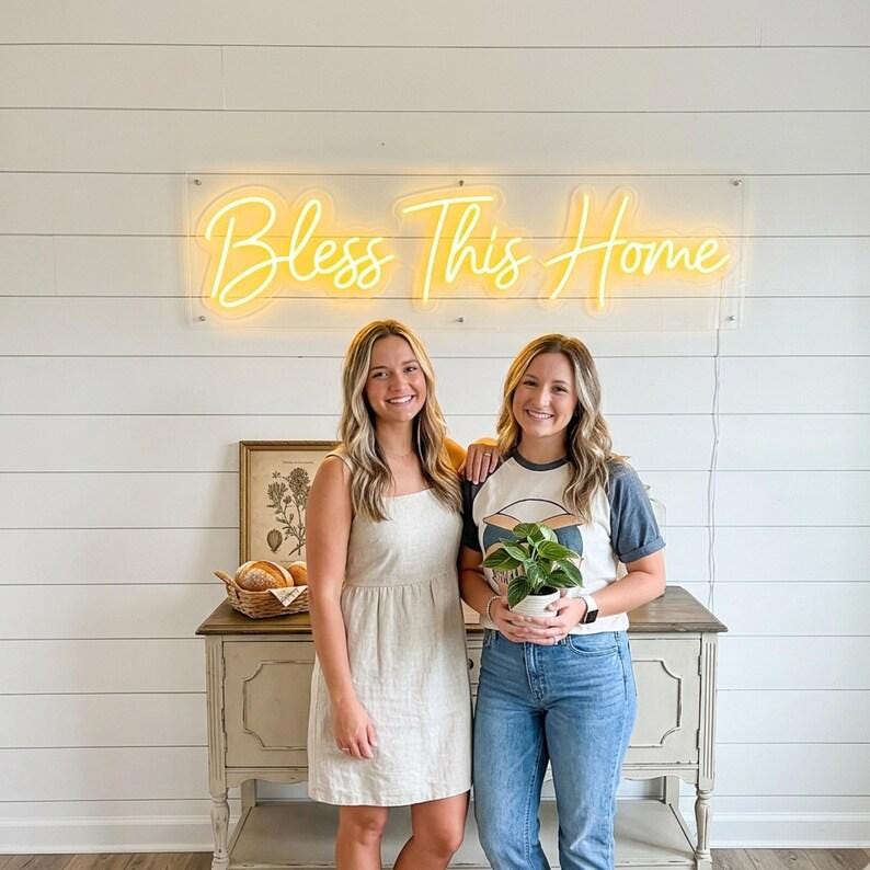 May include: A rectangular neon sign with yellow script lettering that reads Bless This Home, mounted on a clear acrylic backing. The sign is positioned horizontally against a white horizontal plank wall above a wooden console table.