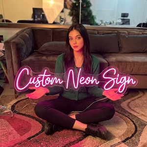 May include: A bright pink neon sign featuring the text Custom Neon Sign in a cursive script font. The sign is mounted on a clear acrylic backing and emits a vibrant glow, illuminating the surrounding area with a distinct pink light.