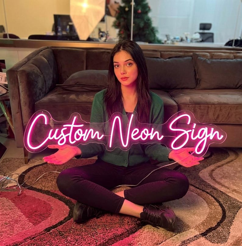 May include: A bright pink neon sign featuring the text Custom Neon Sign in a cursive script font. The sign is mounted on a clear acrylic backing and emits a vibrant glow, illuminating the surrounding area with a distinct pink light.