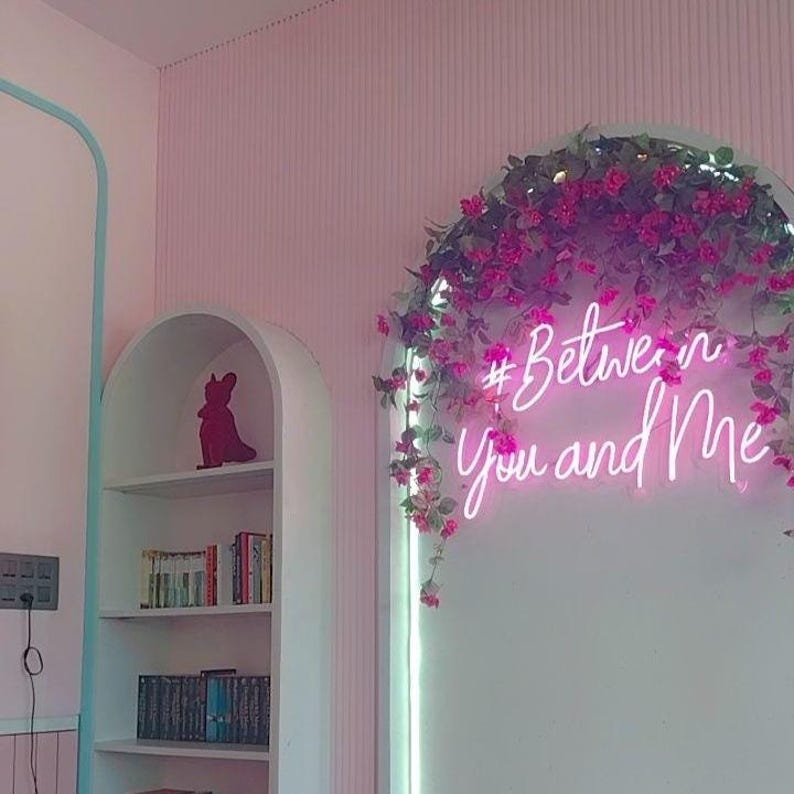 May include: A white arched wall display features a bright pink neon sign reading #Between You and Me, adorned with cascading dark pink floral vines. Beside it, a white arched bookshelf holds books and a small dark pink decorative figurine.