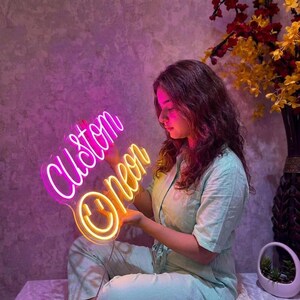 May include: A custom neon sign featuring the word Custom in pink script lettering above the word Neon in yellow script lettering, with a yellow smiley face icon at the bottom. The sign emits a bright glow against a textured wall background.