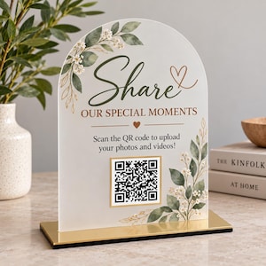 May include: An arched acrylic sign with a gold base featuring green watercolor leaves and white flowers. The text reads Share OUR SPECIAL MOMENTS and Scan the QR code to upload your photos and videos! A central QR code is framed in gold.