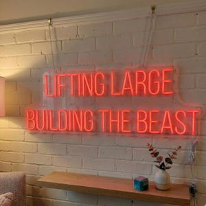 May include: A rectangular neon sign with red lettering mounted on a white brick wall. The sign displays the text LIFTING LARGE on the top line and BUILDING THE BEAST on the bottom line. The sign is suspended by two thin metal chains from the top.