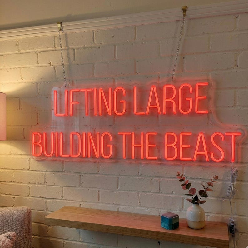 May include: A rectangular neon sign with red lettering mounted on a white brick wall. The sign displays the text LIFTING LARGE on the top line and BUILDING THE BEAST on the bottom line. The sign is suspended by two thin metal chains from the top.