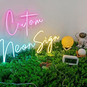 May include: A custom neon sign with pink and yellow cursive lettering that reads "Cutom Neon Sign". The sign is displayed above a patch of green artificial foliage alongside a glowing yellow orb, two small astronaut figurines, and a digital clock.