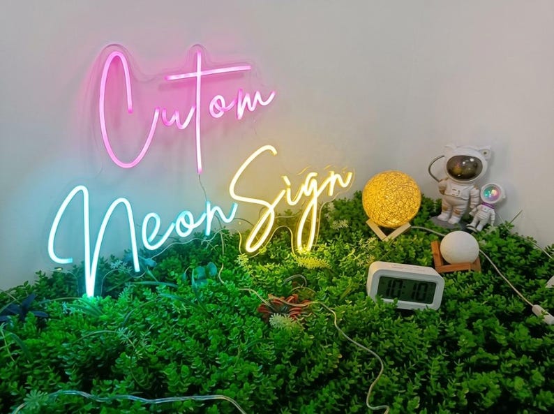 May include: A custom neon sign with pink and yellow cursive lettering that reads "Cutom Neon Sign". The sign is displayed above a patch of green artificial foliage alongside a glowing yellow orb, two small astronaut figurines, and a digital clock.