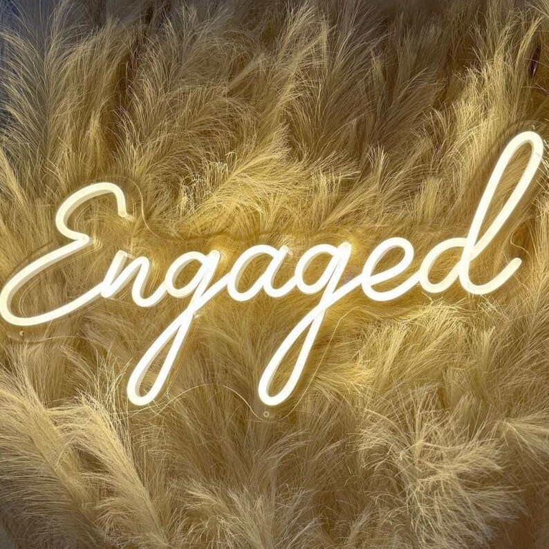 May include: A white neon sign featuring the word Engaged in a cursive script font. The sign is mounted against a backdrop of fluffy, light tan pampas grass plumes that create a soft, textured, and neutral-toned visual display behind the lettering.