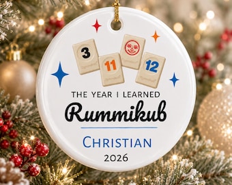 May include: A round white ceramic ornament featuring four Rummikub tiles with the numbers 3, 11, 12, and a sun icon. Text reads: THE YEAR I LEARNED Rummikub, CHRISTIAN, and 2026. The ornament includes a gold hanging string and colorful star accents.