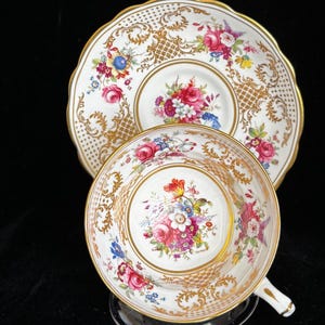May include: A white teacup and saucer set with a floral design. The set features a gold rim and accents, with a detailed pattern of pink, red, and blue flowers. The cup is on a clear stand.