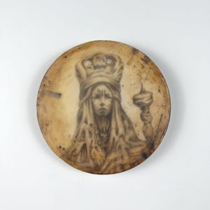 Può includere: Un piatto decorativo circolare di colore marrone chiaro dall'aspetto usurato, caratterizzato da un'illustrazione color seppia di una figura coronata con capelli lunghi e segni sul viso, che tiene in mano uno scettro. La superficie presenta graffi.