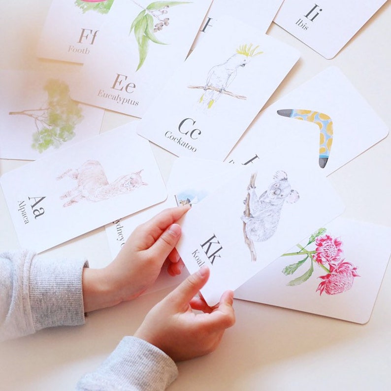 Puede incluir: Un conjunto de tarjetas de memoria del alfabeto con animales y plantas australianas. Las tarjetas son blancas con letras negras e ilustraciones coloridas. Las tarjetas incluyen las letras A a K, con palabras como "Alpaca", "Cacat&uacute;a", "Koala" y "Eucalipto".