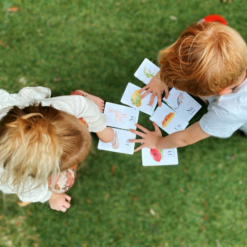 Puede incluir: Dos ni&ntilde;os juegan con tarjetas de letras en un c&eacute;sped verde. Las tarjetas muestran ilustraciones coloridas de animales y las letras Aa, Hh, Ll, Uu, Nn y Ff.